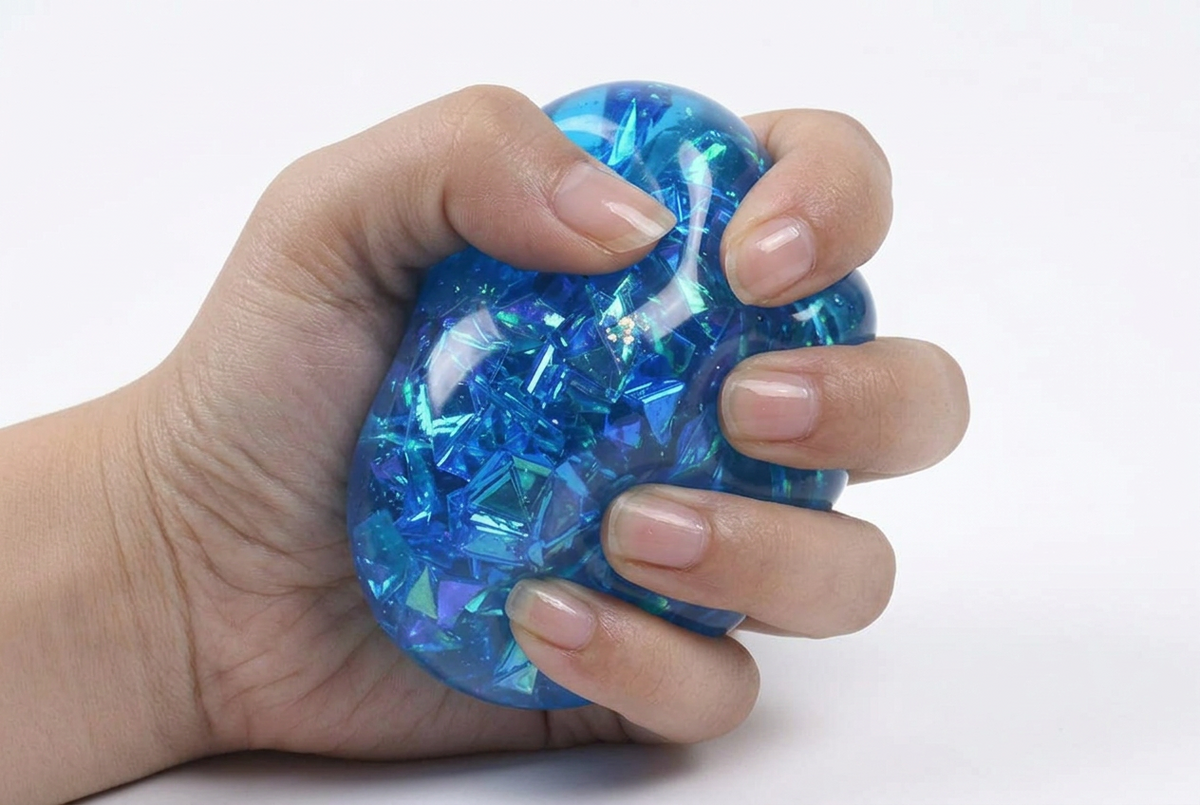 A hand squeezing a translucent blue stress ball filled with iridescent blue and green reflective pieces, shown against a plain white background.
