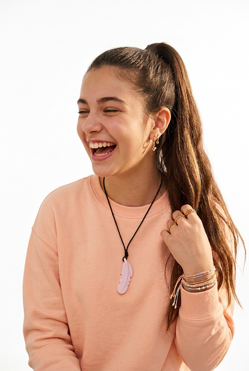 A young woman with a high ponytail is laughing, wearing a peach-colored sweatshirt and a black cord necklace with a light pink feather chew pendant. She is set against a plain white background.